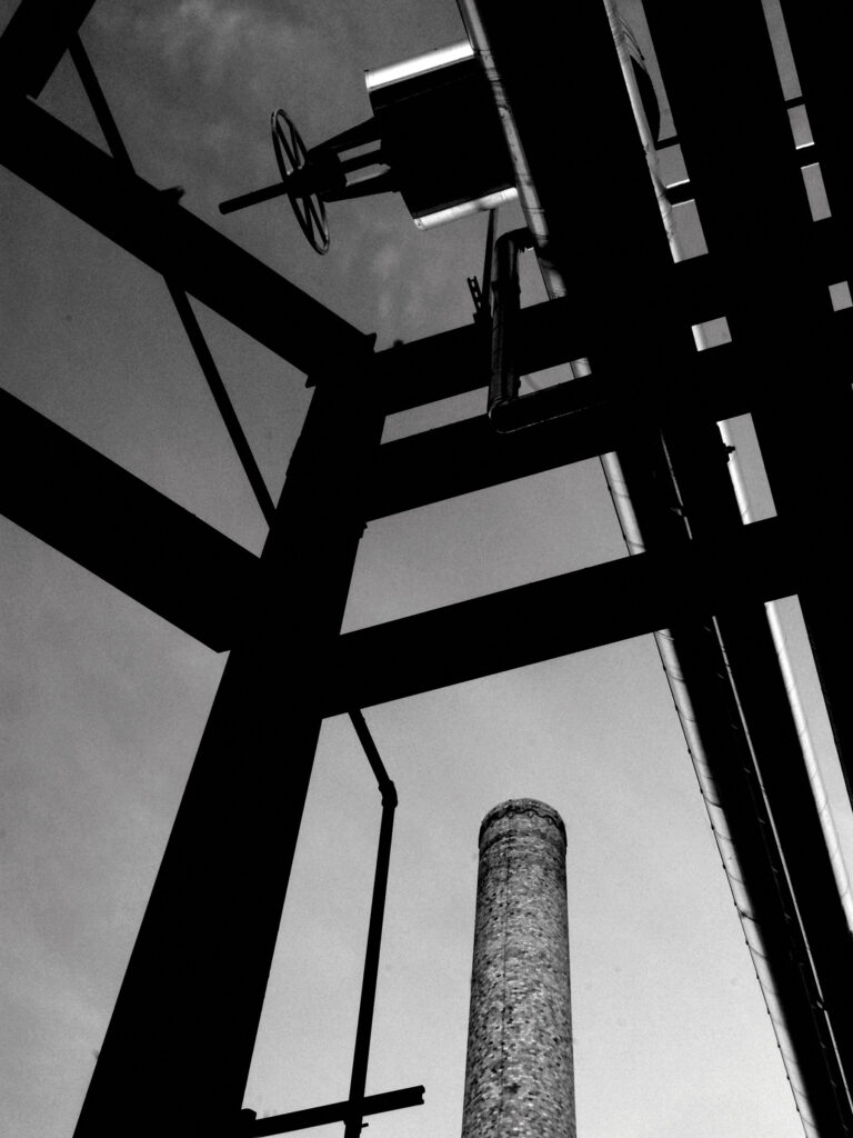 A black-and-white photo looking up at the chimney stack of a power plant framed by several beams