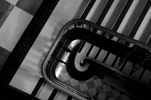 A black and white photo looking down the levels of a grand staircase. There is a checkered floor.