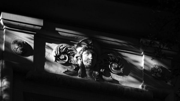 A black and white photo of a wall in shadow. The light is hitting a sculpture detail of a Classical Greek face