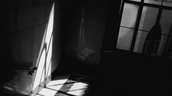 A black and white photo of a staircase landing in shadow. The light from a wind fours a triangular shape on the wall.