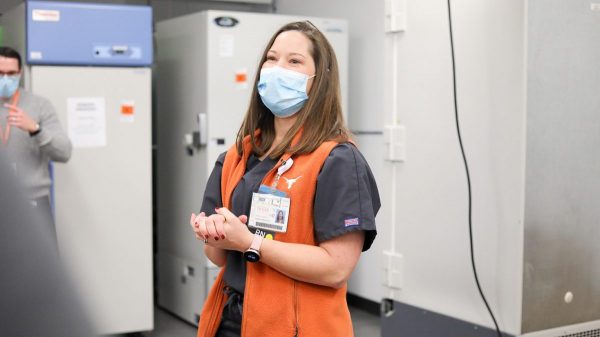 A woman wearing a hospital mask and an orange vest stands before laboratory freezers.