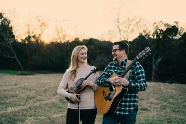 A woman holding a violin and a man holding an acoustic guitar pose together outside in front of a wooded area.