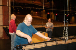 A man uses a measuring tape on a stage as equipment and backdrops are brought in.