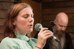 A woman sings into a microphone as a man in the background plays guitar.