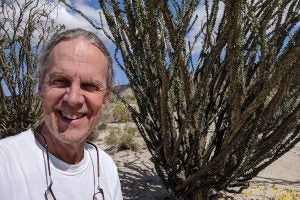 A man in a white T-shirt stands before a large succulent in a desert landscape.