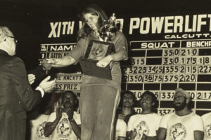 Vintage photo of Jan Todd accepting a powerlifting award with the United States men's team behind her